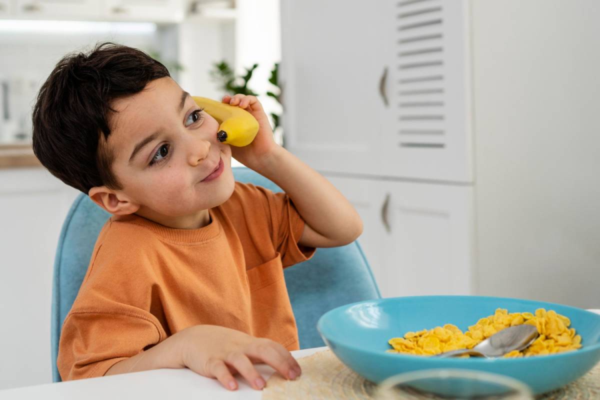 Little boy sitting at the dining table with a bowl of cereal and a banana, showing how children learn healthy food choices through play and routine