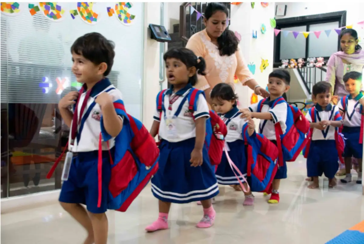 Screenshot 2026-04-27 162937 Preschool children in uniforms walking in a line with backpacks, guided by a teacher inside a colorful daycare classroom