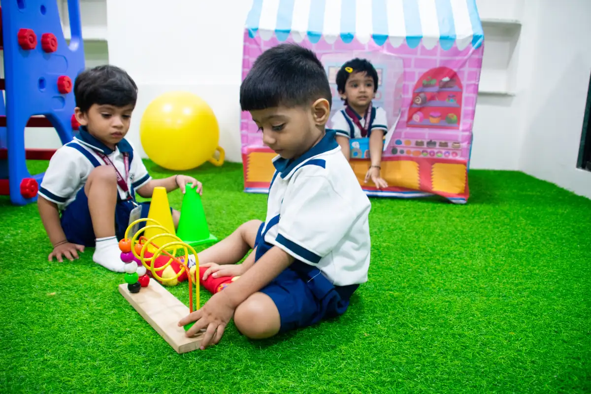Children playing with colourful toys and building blocks on green artificial grass in a preschool classroom environment.