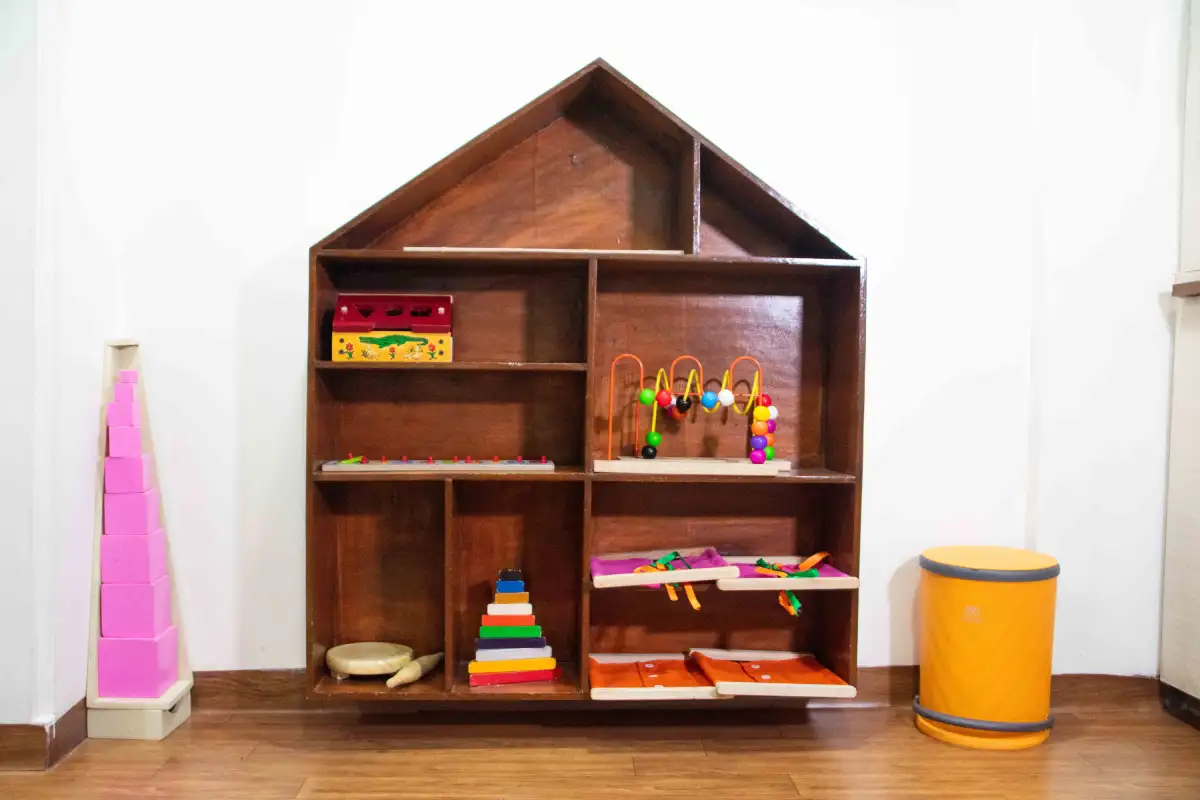 ooden house-shaped Montessori toy shelf with multiple compartments holding educational toys, including stacking rings, bead maze, shape sorter, and activity materials, placed on a wooden floor with a pink tower toy on the left and a yellow storage bin on the right.